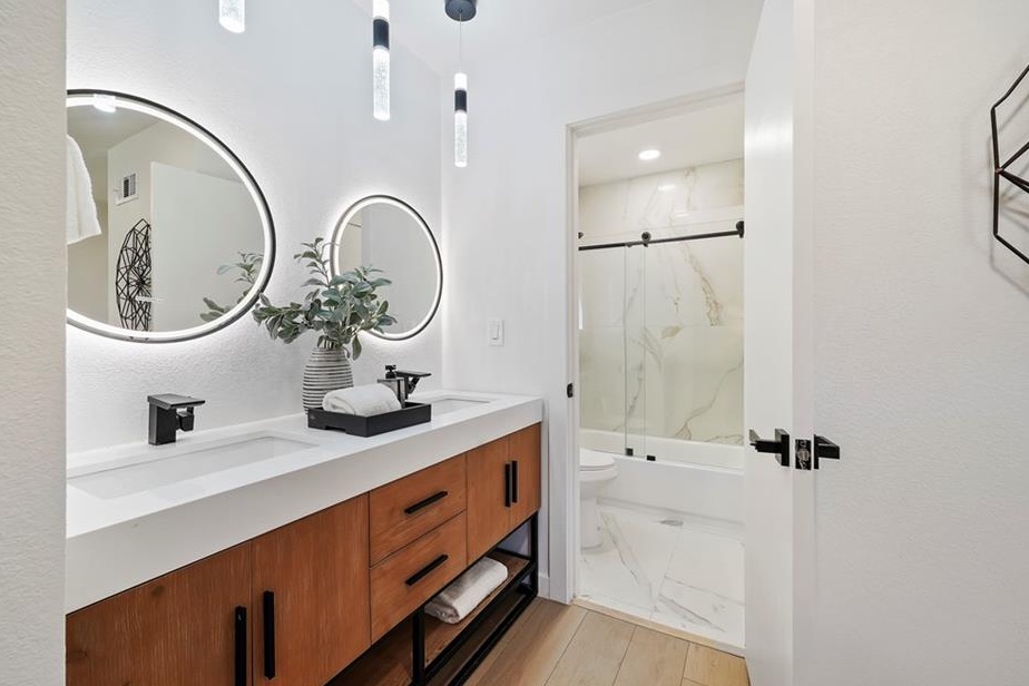 Remodeled bathroom with a wood double vanity, backlit oval mirrors, pendant lighting, and a marble-look tiled shower and tub