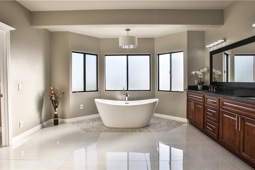 Remodeled primary bathroom with a freestanding soaker tub set into a windowed bay, a double wood vanity, and a polished stone floor
